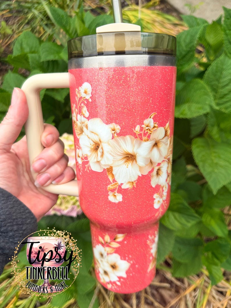 K&ouml;nnte beinhalten: Ein korallenfarbener Becher mit Glitzer-Finish, verziert mit einem wei&szlig;en Blumenmuster, einem beigen Henkel und einem dunkel durchscheinenden Deckel mit Strohhalm. Der Becher zeigt ein Logo mit dem Text Tipsy Tinkerbell Tumblers & More.