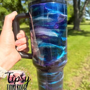 May include: A large, insulated tumbler with a black handle and lid. The tumbler features a swirling design of blue, purple, and white, with glitter accents. The text "Tipsy Tinkerbell Tumblers & More" is visible.