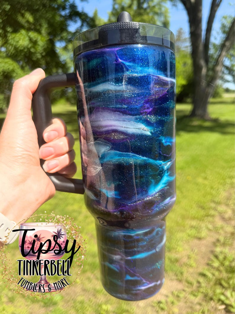 May include: A large, insulated tumbler with a black handle and lid. The tumbler features a swirling design of blue, purple, and white, with glitter accents. The text "Tipsy Tinkerbell Tumblers & More" is visible.