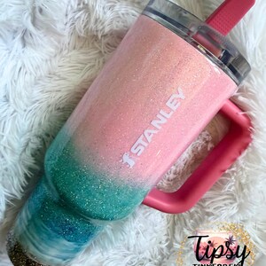 May include: A Stanley tumbler featuring a glittery ombre finish that transitions from light pink at the top to teal and dark blue at the base. The cup has a pink handle, a clear lid, and a pink straw. The word STANLEY is printed in white on the side.