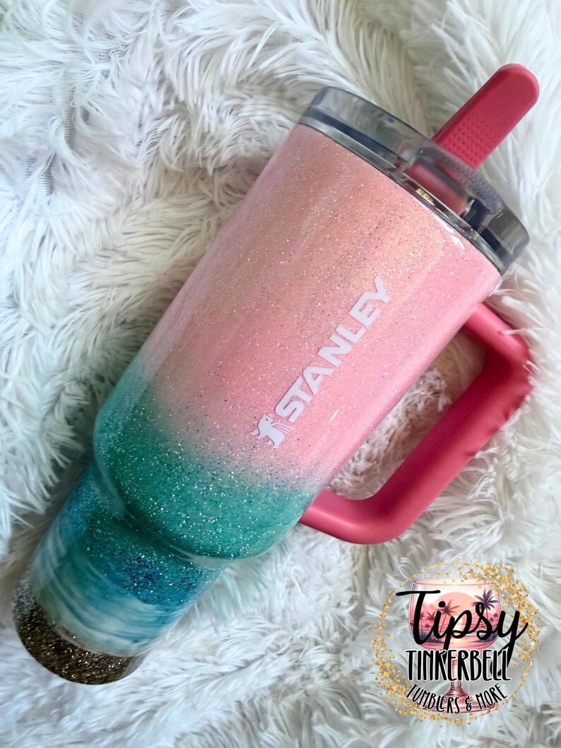 May include: A Stanley tumbler featuring a glittery ombre finish that transitions from light pink at the top to teal and dark blue at the base. The cup has a pink handle, a clear lid, and a pink straw. The word STANLEY is printed in white on the side.