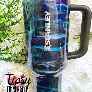 May include: A Stanley tumbler with a black handle and lid features a swirling design in shades of blue, teal, purple, and silver. The tumbler has the Stanley logo in silver. The bottom of the tumbler has the text "Tipsy Tinkerbell Tumblers & More".