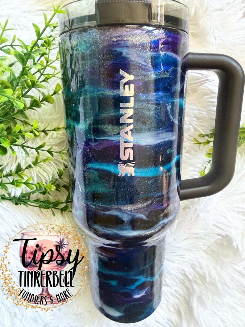 May include: A Stanley tumbler with a black handle and lid features a swirling design in shades of blue, teal, purple, and silver. The tumbler has the Stanley logo in silver. The bottom of the tumbler has the text "Tipsy Tinkerbell Tumblers & More".