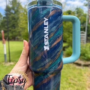 Puede incluir: Un vaso Stanley azul y verde azulado con asa. El vaso tiene un dise&ntilde;o de purpurina arremolinada con detalles dorados. La palabra "STANLEY" est&aacute; impresa en blanco. El asa es de color azul claro.