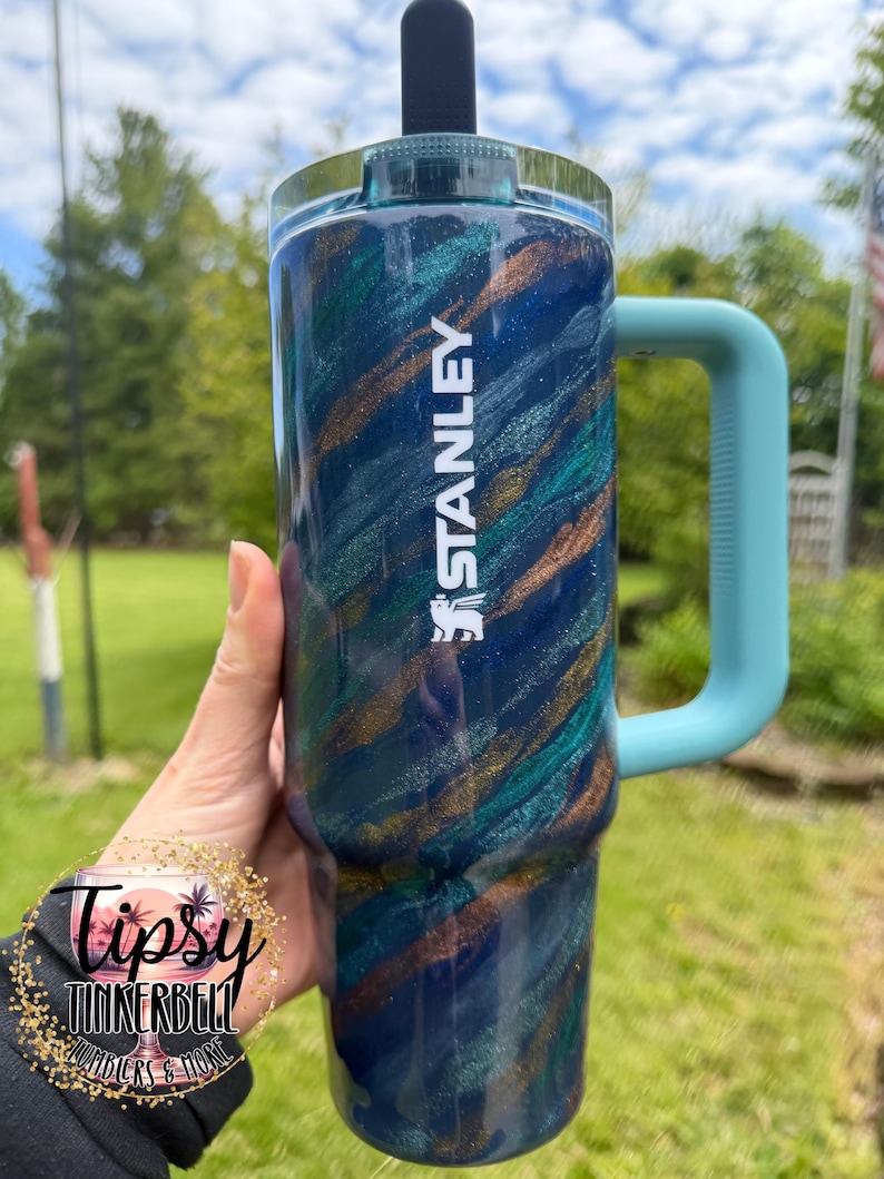 Puede incluir: Un vaso Stanley azul y verde azulado con asa. El vaso tiene un dise&ntilde;o de purpurina arremolinada con detalles dorados. La palabra "STANLEY" est&aacute; impresa en blanco. El asa es de color azul claro.
