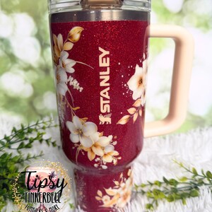 Puede incluir: Un vaso Stanley rojo brillante con un dise&ntilde;o floral. El vaso presenta flores blancas y amarillas y el logotipo de Stanley. Tiene un asa rosa claro y una tapa transparente.