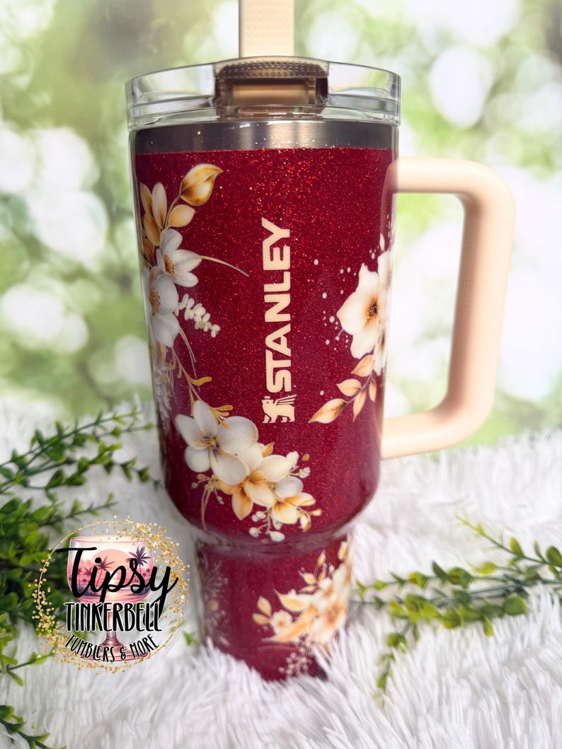 Puede incluir: Un vaso Stanley rojo brillante con un dise&ntilde;o floral. El vaso presenta flores blancas y amarillas y el logotipo de Stanley. Tiene un asa rosa claro y una tapa transparente.
