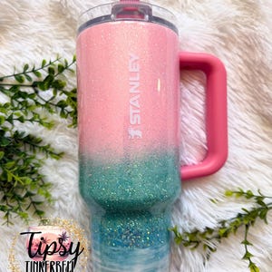 May include: A Stanley tumbler featuring a pink and teal glitter ombre finish that transitions into a beach-themed design with blue water, white waves, and a sandy base. The cup includes a pink handle, a clear lid, and a pink straw.