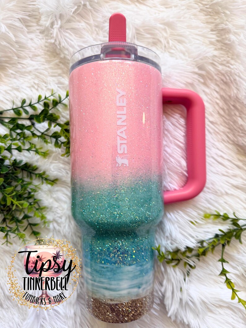 May include: A Stanley tumbler featuring a pink and teal glitter ombre finish that transitions into a beach-themed design with blue water, white waves, and a sandy base. The cup includes a pink handle, a clear lid, and a pink straw.