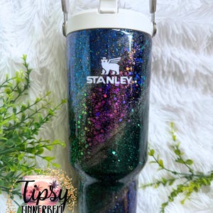 May include: A Stanley tumbler featuring a dark, glittery finish with swirling shades of blue, purple, and green. The cup has a white lid with a handle and the white Stanley logo printed on the front. A circular logo reads Tipsy Tinkerbell.