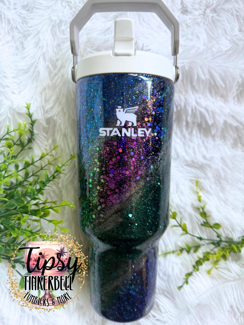 May include: A Stanley tumbler featuring a dark, glittery finish with swirling shades of blue, purple, and green. The cup has a white lid with a handle and the white Stanley logo printed on the front. A circular logo reads Tipsy Tinkerbell.