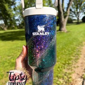 May include: A Stanley brand tumbler featuring a dark blue to green gradient finish covered in iridescent glitter. The tumbler has a white lid with a handle and a straw opening. The white Stanley logo is printed on the upper front of the cup.