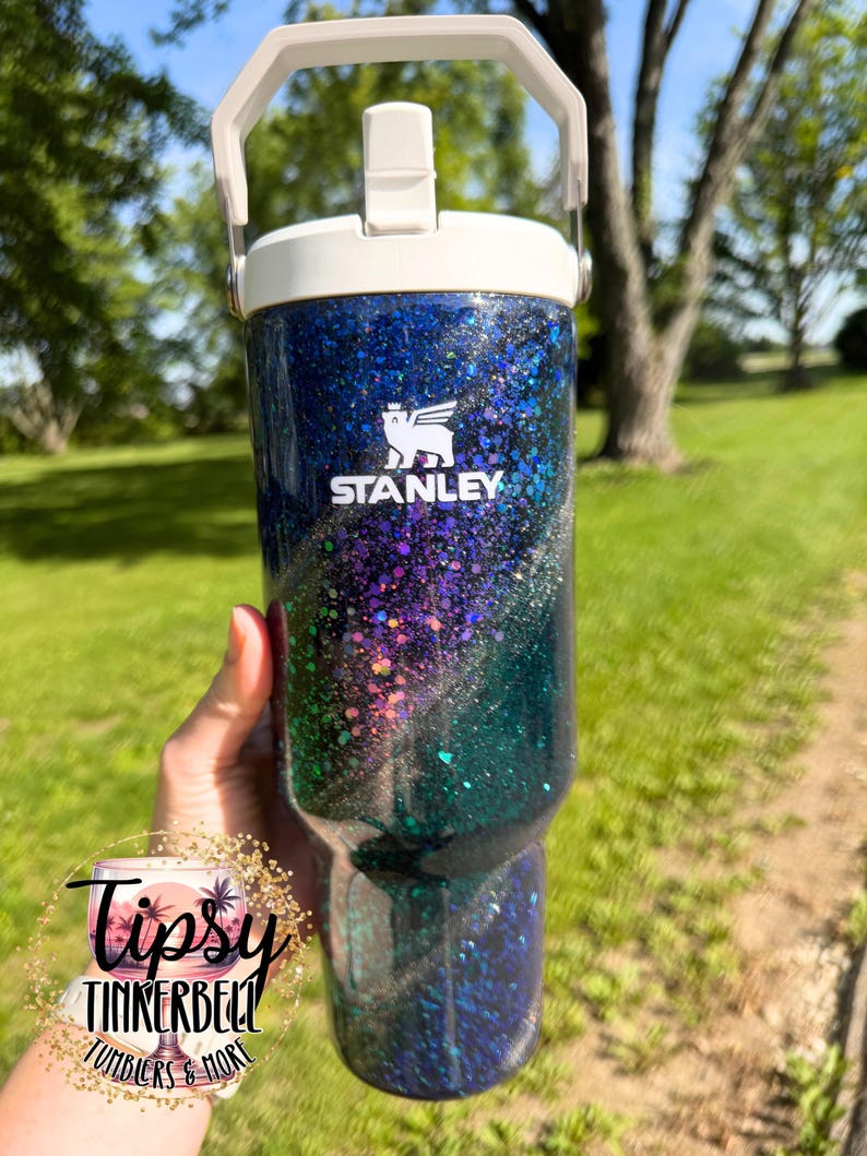 May include: A Stanley brand tumbler featuring a dark blue to green gradient finish covered in iridescent glitter. The tumbler has a white lid with a handle and a straw opening. The white Stanley logo is printed on the upper front of the cup.