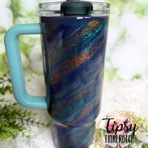 Puede incluir: Un vaso con un dise&ntilde;o en espiral azul, verde azulado y dorado. Tiene un asa azul claro y una tapa gris oscuro con cierre abatible. El vaso tiene el texto "Tipsy Tinkerbell Tumblers & More" en un logotipo circular.
