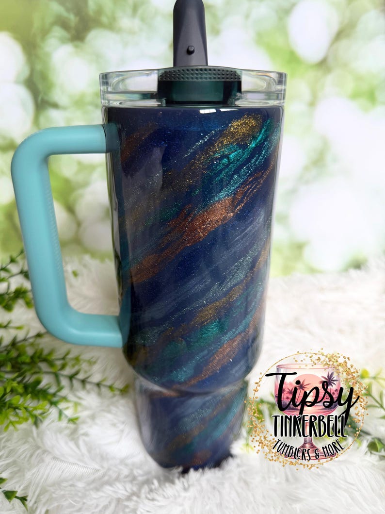 Puede incluir: Un vaso con un dise&ntilde;o en espiral azul, verde azulado y dorado. Tiene un asa azul claro y una tapa gris oscuro con cierre abatible. El vaso tiene el texto "Tipsy Tinkerbell Tumblers & More" en un logotipo circular.