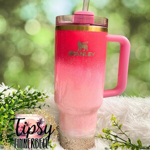 May include: A pink and white ombre Stanley tumbler with a pink handle and lid. The tumbler has a gold rim and a straw. The bottom of the tumbler has a sand-like texture. The tumbler has the Stanley logo on the front.