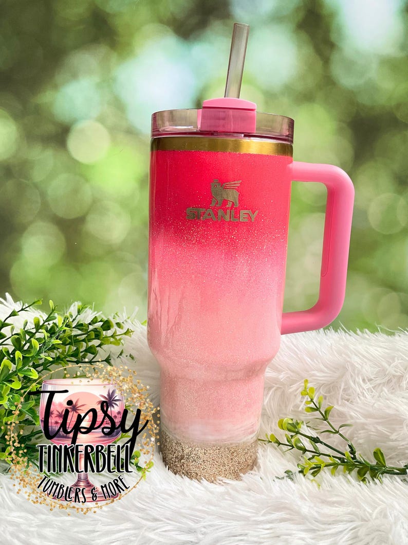 May include: A pink and white ombre Stanley tumbler with a pink handle and lid. The tumbler has a gold rim and a straw. The bottom of the tumbler has a sand-like texture. The tumbler has the Stanley logo on the front.
