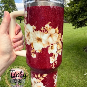 Puede incluir: Un vaso grande con purpurina roja con un asa y tapa color crema. El vaso est&aacute; decorado con dise&ntilde;os florales blancos y dorados. El vaso tiene el texto "Tipsy Tinkerbell Tumblers & More" en un logotipo circular.