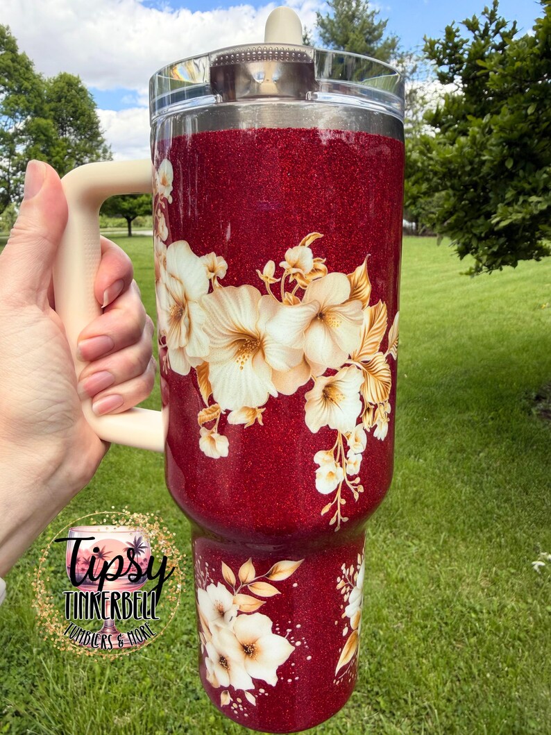 Puede incluir: Un vaso grande con purpurina roja con un asa y tapa color crema. El vaso est&aacute; decorado con dise&ntilde;os florales blancos y dorados. El vaso tiene el texto "Tipsy Tinkerbell Tumblers & More" en un logotipo circular.