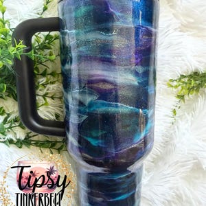May include: A large, insulated tumbler with a black handle and lid. The tumbler features a swirling design in shades of blue, teal, purple, and silver. The text "Tipsy Tinkerbell Tumblers & More" is visible on the bottom left.