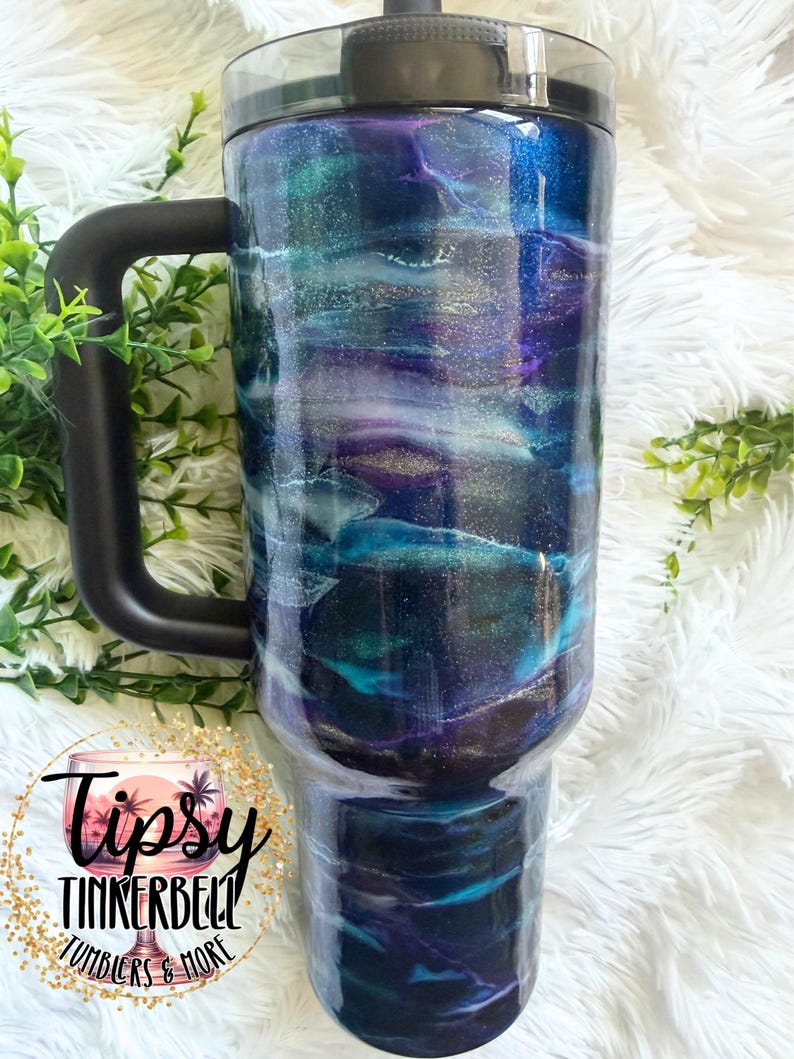 May include: A large, insulated tumbler with a black handle and lid. The tumbler features a swirling design in shades of blue, teal, purple, and silver. The text "Tipsy Tinkerbell Tumblers & More" is visible on the bottom left.