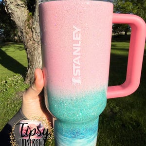 May include: A Stanley tumbler featuring a pink glitter top section that transitions into a teal glitter middle, followed by a blue and white ocean wave design at the base, finished with a sandy texture at the bottom and a pink handle and lid.