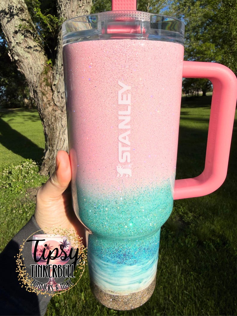 May include: A Stanley tumbler featuring a pink glitter top section that transitions into a teal glitter middle, followed by a blue and white ocean wave design at the base, finished with a sandy texture at the bottom and a pink handle and lid.