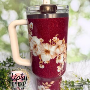 Puede incluir: Un vaso rojo brillante con asa y tapa color crema. El vaso presenta un dise&ntilde;o floral con flores blancas y beige. El texto "Tipsy Tinkerbell Tumblers & More" es visible en la parte inferior izquierda.