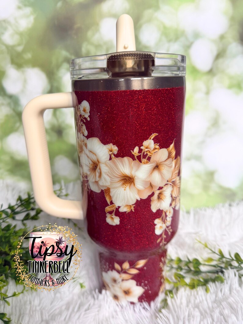 Puede incluir: Un vaso rojo brillante con asa y tapa color crema. El vaso presenta un dise&ntilde;o floral con flores blancas y beige. El texto "Tipsy Tinkerbell Tumblers & More" es visible en la parte inferior izquierda.
