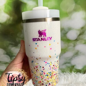 Puede incluir: Vaso blanco con purpurina y dise&ntilde;o de confeti colorido. El vaso presenta el logotipo "STANLEY" en morado y una pajita verde claro. La parte inferior del vaso est&aacute; cubierta de confeti colorido.