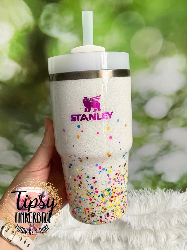 Puede incluir: Vaso blanco con purpurina y dise&ntilde;o de confeti colorido. El vaso presenta el logotipo "STANLEY" en morado y una pajita verde claro. La parte inferior del vaso est&aacute; cubierta de confeti colorido.
