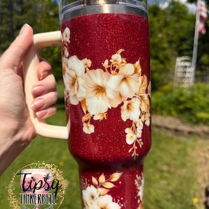Puede incluir: Un vaso rojo brillante con un asa color crema y un dise&ntilde;o floral. El vaso presenta una tapa transparente y un patr&oacute;n floral decorativo en crema y dorado. El vaso tiene el texto "Tipsy Tinkerbell Tumblers & More".