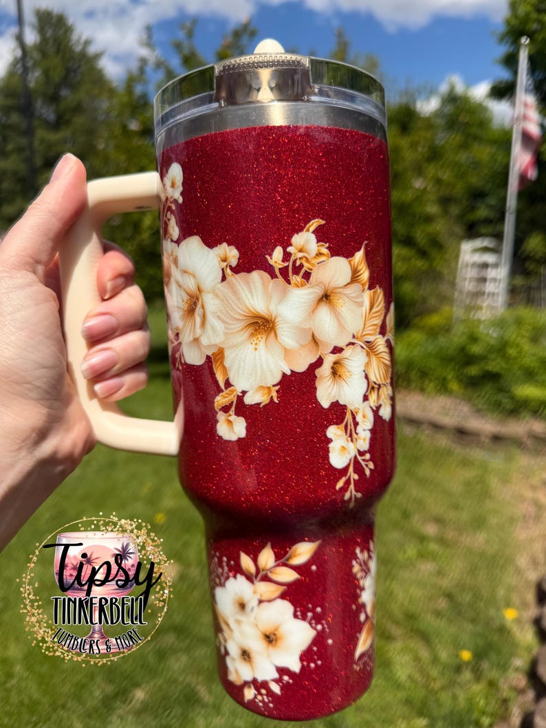 Puede incluir: Un vaso rojo brillante con un asa color crema y un dise&ntilde;o floral. El vaso presenta una tapa transparente y un patr&oacute;n floral decorativo en crema y dorado. El vaso tiene el texto "Tipsy Tinkerbell Tumblers & More".