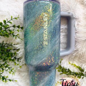 May include: A Stanley tumbler with a blue and white glitter design. The tumbler has a handle and a clear lid. The word "STANLEY" is visible on the side. The tumbler is decorated with iridescent glitter and the words "Tipsy Tinkerbell Tumblers & More".