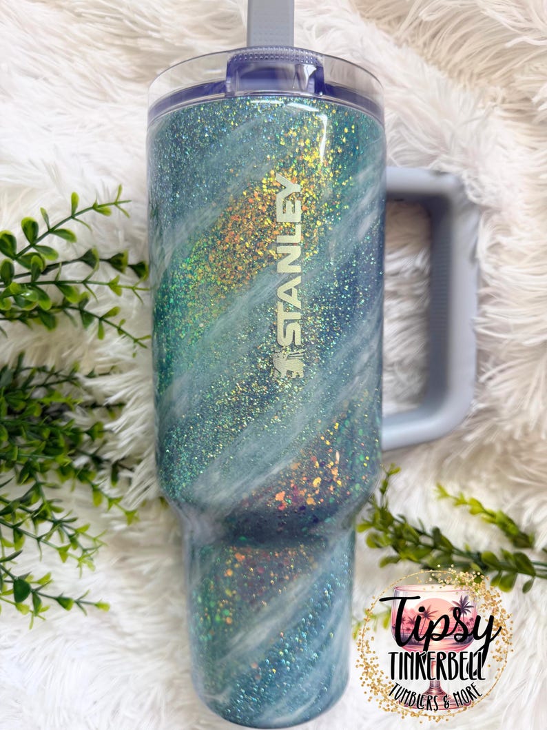 May include: A Stanley tumbler with a blue and white glitter design. The tumbler has a handle and a clear lid. The word "STANLEY" is visible on the side. The tumbler is decorated with iridescent glitter and the words "Tipsy Tinkerbell Tumblers & More".