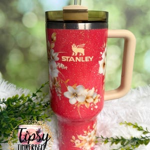 K&ouml;nnte beinhalten: Ein roter Stanley-Becher mit Glitzereffekt und einem Blumenmuster aus wei&szlig;en und goldenen Bl&uuml;ten. Der Becher hat einen beigen Henkel, einen dunkel durchscheinenden Deckel und einen transparenten Strohhalm. Das Stanley-Logo ist goldfarben aufgedruckt.