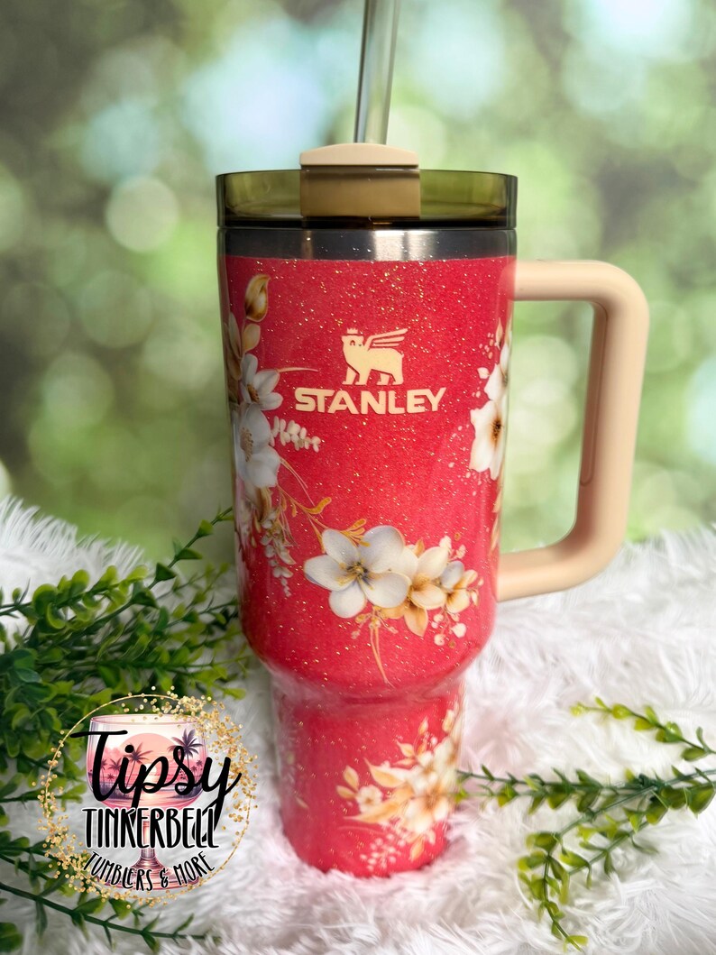 K&ouml;nnte beinhalten: Ein roter Stanley-Becher mit Glitzereffekt und einem Blumenmuster aus wei&szlig;en und goldenen Bl&uuml;ten. Der Becher hat einen beigen Henkel, einen dunkel durchscheinenden Deckel und einen transparenten Strohhalm. Das Stanley-Logo ist goldfarben aufgedruckt.