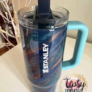 Puede incluir: Un vaso Stanley con un dise&ntilde;o de remolino azul, verde azulado y dorado. El vaso tiene una tapa transparente, un asa azul y el logotipo de Stanley en blanco. El vaso tambi&eacute;n tiene el texto "Tipsy Tinkerbell Tumblers & More".