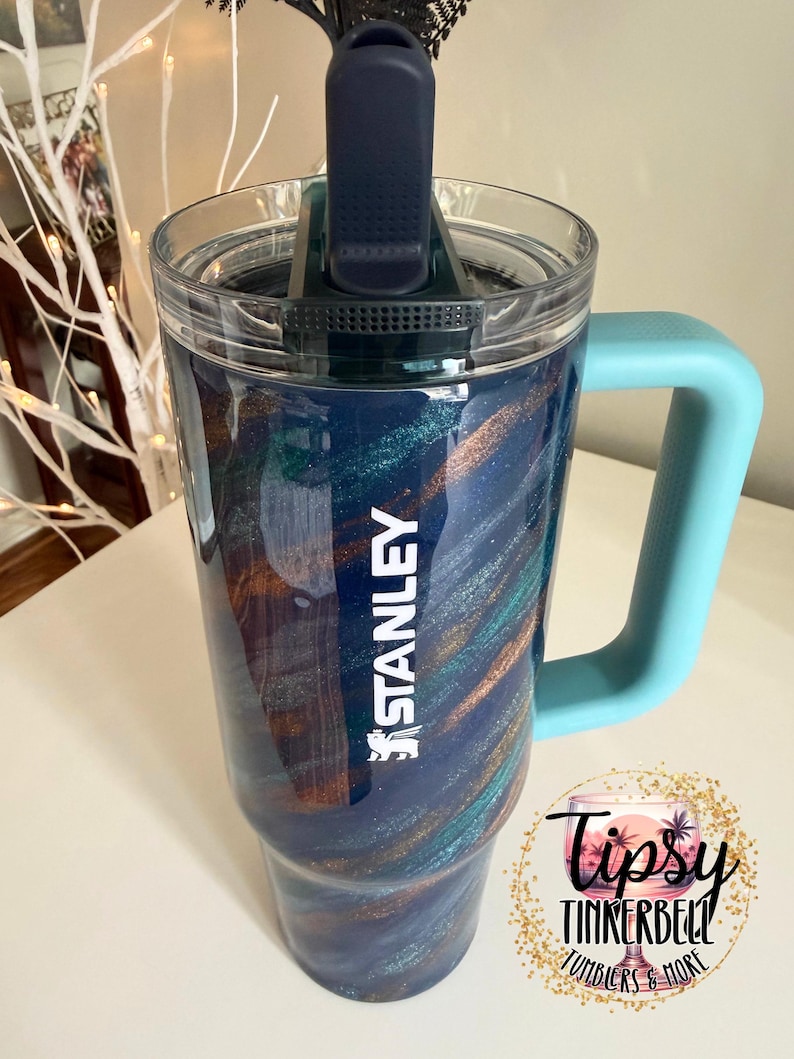 Puede incluir: Un vaso Stanley con un dise&ntilde;o de remolino azul, verde azulado y dorado. El vaso tiene una tapa transparente, un asa azul y el logotipo de Stanley en blanco. El vaso tambi&eacute;n tiene el texto "Tipsy Tinkerbell Tumblers & More".
