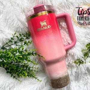 May include: A pink and gold Stanley tumbler with a handle. The tumbler has a gradient design, transitioning from a bright pink at the top to a lighter pink and gold glitter at the bottom. The Stanley logo is visible.