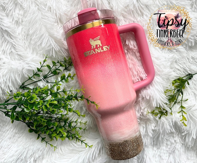 May include: A pink and gold Stanley tumbler with a handle. The tumbler has a gradient design, transitioning from a bright pink at the top to a lighter pink and gold glitter at the bottom. The Stanley logo is visible.