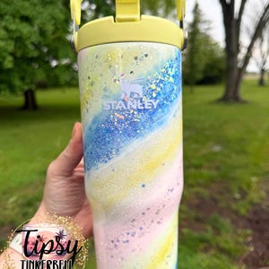 May include: A Stanley tumbler with a pastel swirl design in yellow, pink, and blue, with glitter accents. The tumbler has a yellow handle and lid. The Stanley logo is visible. The tumbler is a drinkware item.