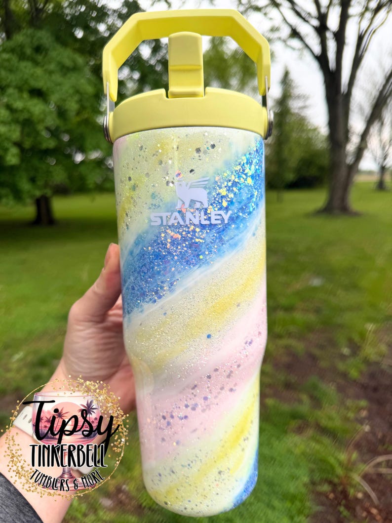 May include: A Stanley tumbler with a pastel swirl design in yellow, pink, and blue, with glitter accents. The tumbler has a yellow handle and lid. The Stanley logo is visible. The tumbler is a drinkware item.