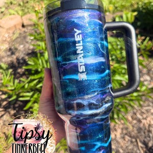 May include: A Stanley tumbler with a black handle and lid. The tumbler features a swirling design of blue, teal, and purple glitter, with the Stanley logo in white. The tumbler is held against a background of green foliage.