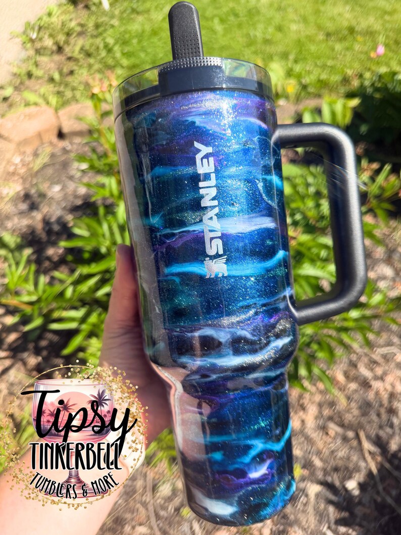 May include: A Stanley tumbler with a black handle and lid. The tumbler features a swirling design of blue, teal, and purple glitter, with the Stanley logo in white. The tumbler is held against a background of green foliage.