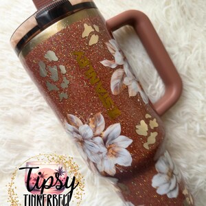 May include: A copper-coloured glitter tumbler with a handle and a straw, featuring a floral pattern of white flowers and gold leopard print spots. The word STANLEY is printed vertically in gold lettering on the side of the cup.