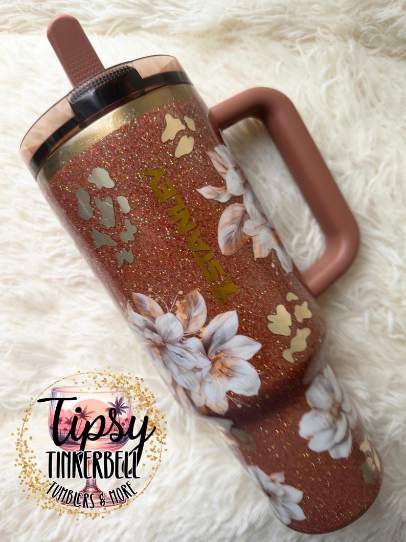 May include: A copper-coloured glitter tumbler with a handle and a straw, featuring a floral pattern of white flowers and gold leopard print spots. The word STANLEY is printed vertically in gold lettering on the side of the cup.