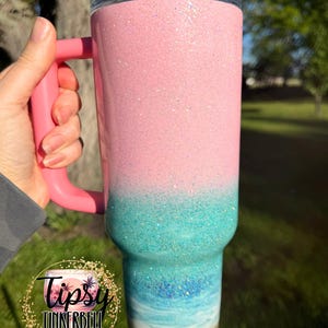 May include: A tall, insulated tumbler with a handle and a clear lid featuring a pink straw. The exterior has a glittery finish that transitions from a solid light pink at the top to a turquoise and blue ocean wave design at the base.