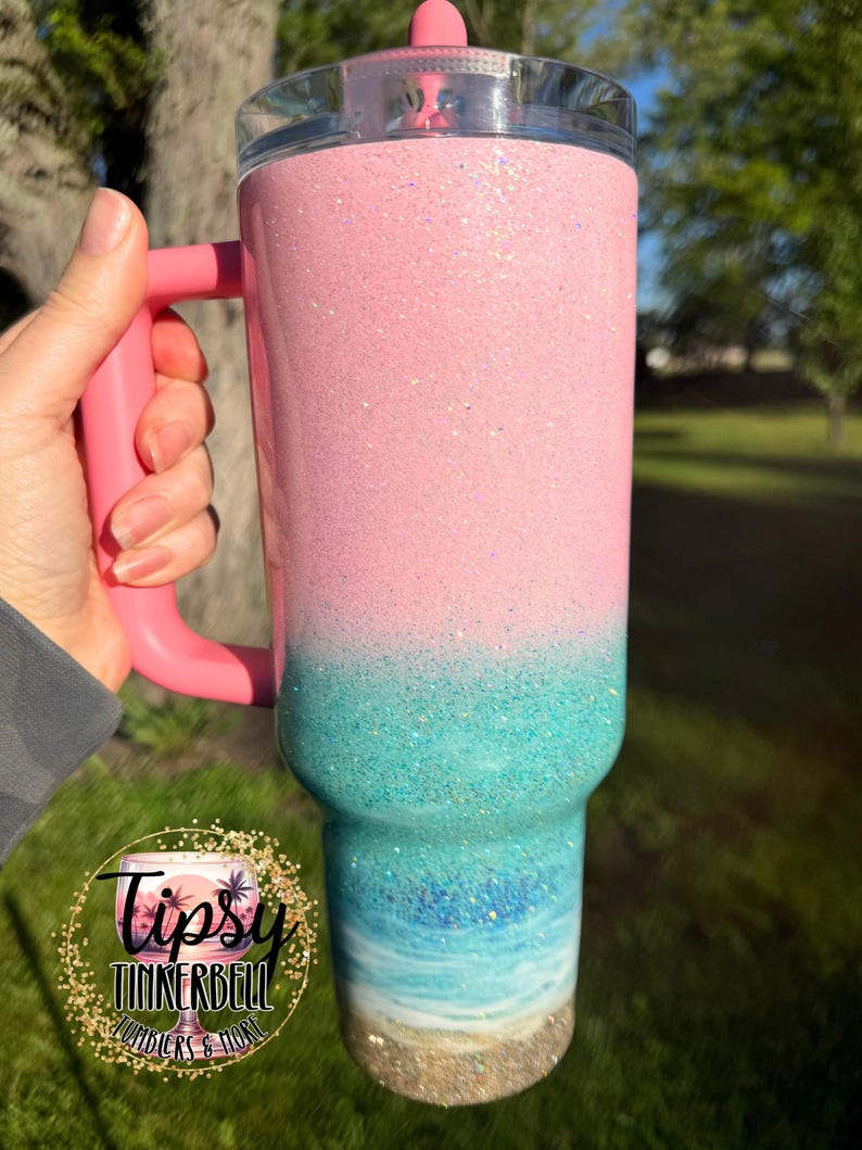 May include: A tall, insulated tumbler with a handle and a clear lid featuring a pink straw. The exterior has a glittery finish that transitions from a solid light pink at the top to a turquoise and blue ocean wave design at the base.