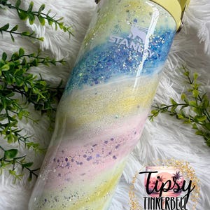 May include: A Stanley tumbler with a pastel swirl design in shades of yellow, blue, pink, and white, with glitter accents. The tumbler has a yellow handle and lid. The text "Stanley" is visible on the side. The bottom right corner has the text "Tipsy Tinkerbell Tumblers & More".